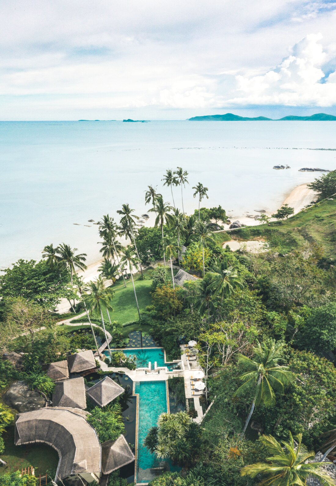 Aerial view of Kamalaya's thatched pavilions and pools terraced into the hillside above a quiet cove, Koh Samui