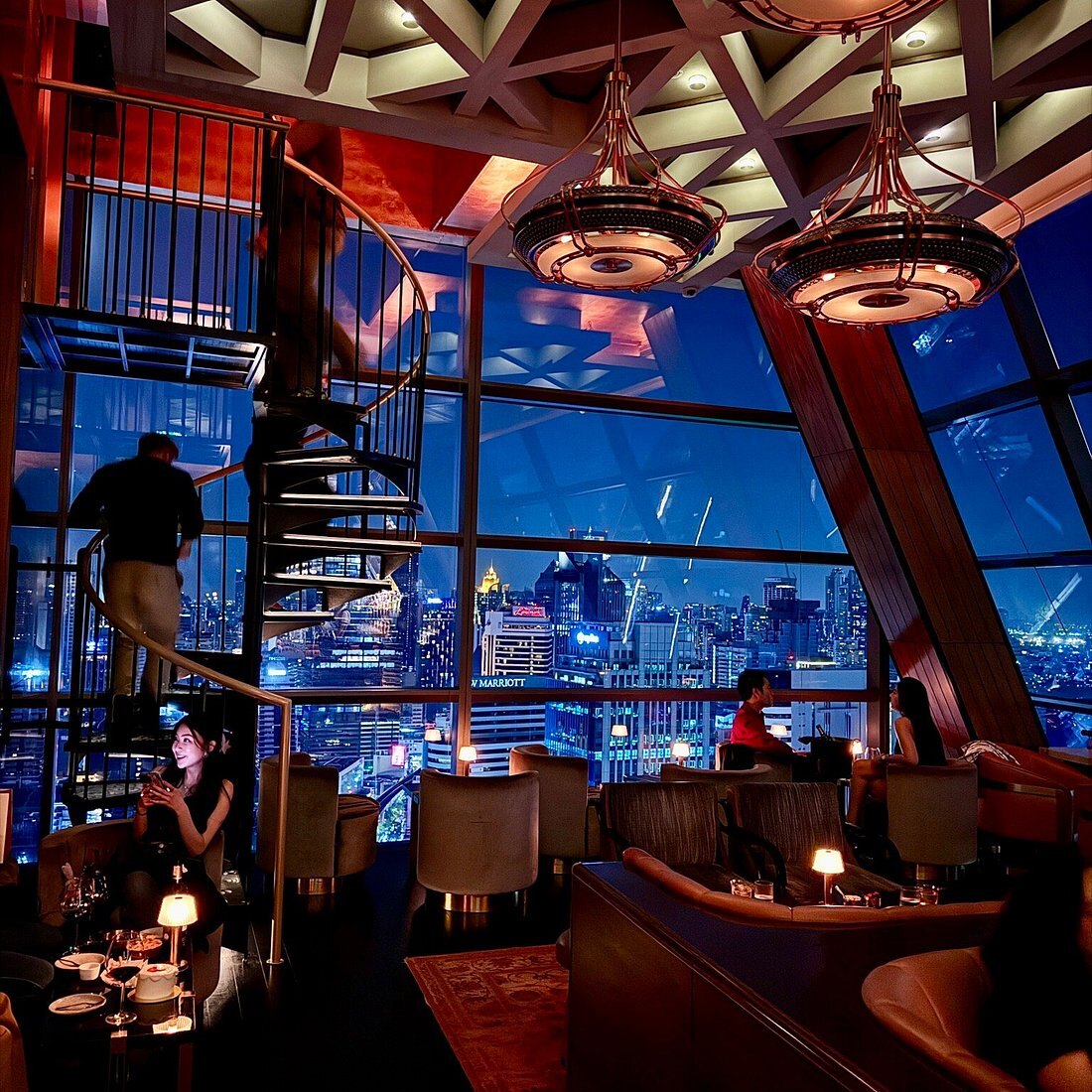 Rosewood Bangkok's bar at night, a spiral staircase between levels and the lit city through full-height glass