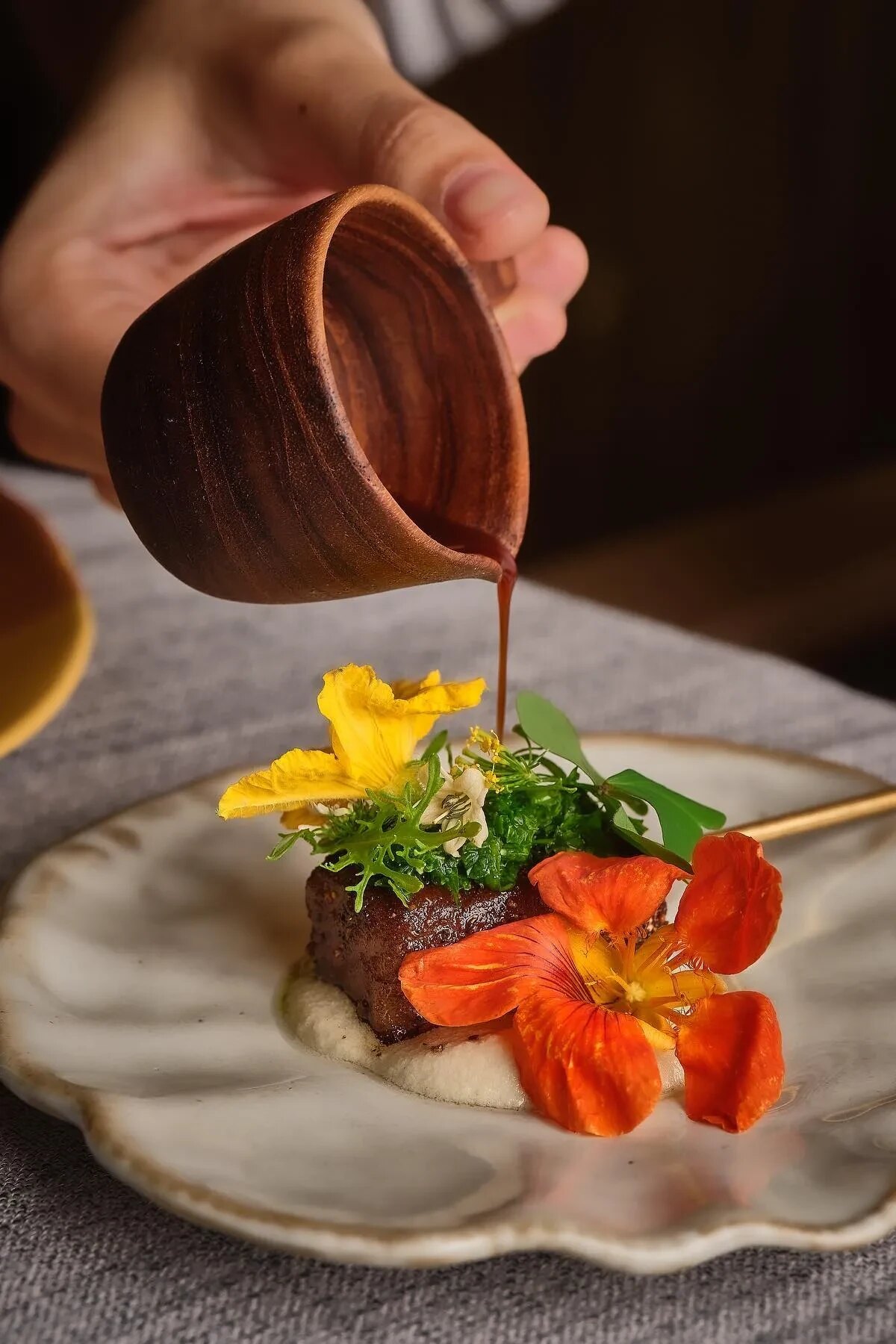 A sauce poured from a turned wooden jug over a plated dish dressed with nasturtium flowers